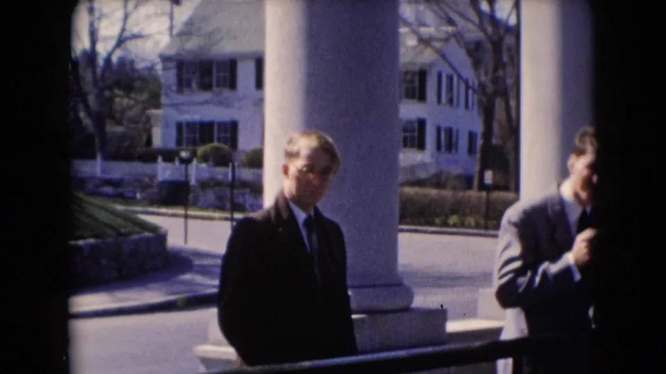A suited man stands near white porch columns, with another man partly cut off at the edge of the frame.