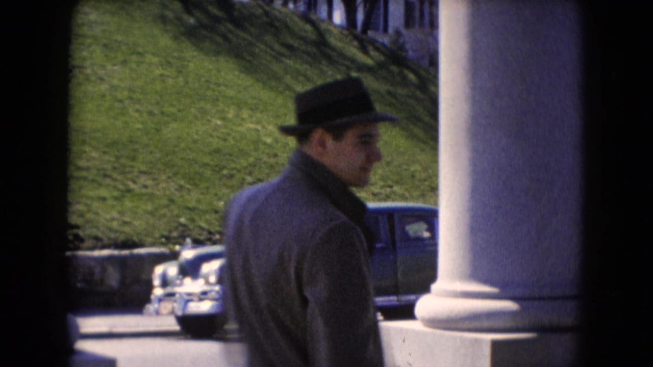 A man wearing a fedora and overcoat stands by a porch column, with a rounded blue car visible on the street behind him.