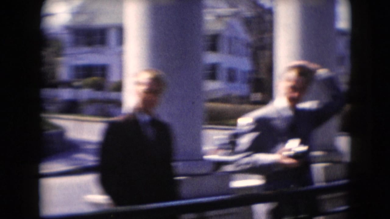 Two suited men appear softly focused near the white porch columns, with one raising a hand while holding an object.
