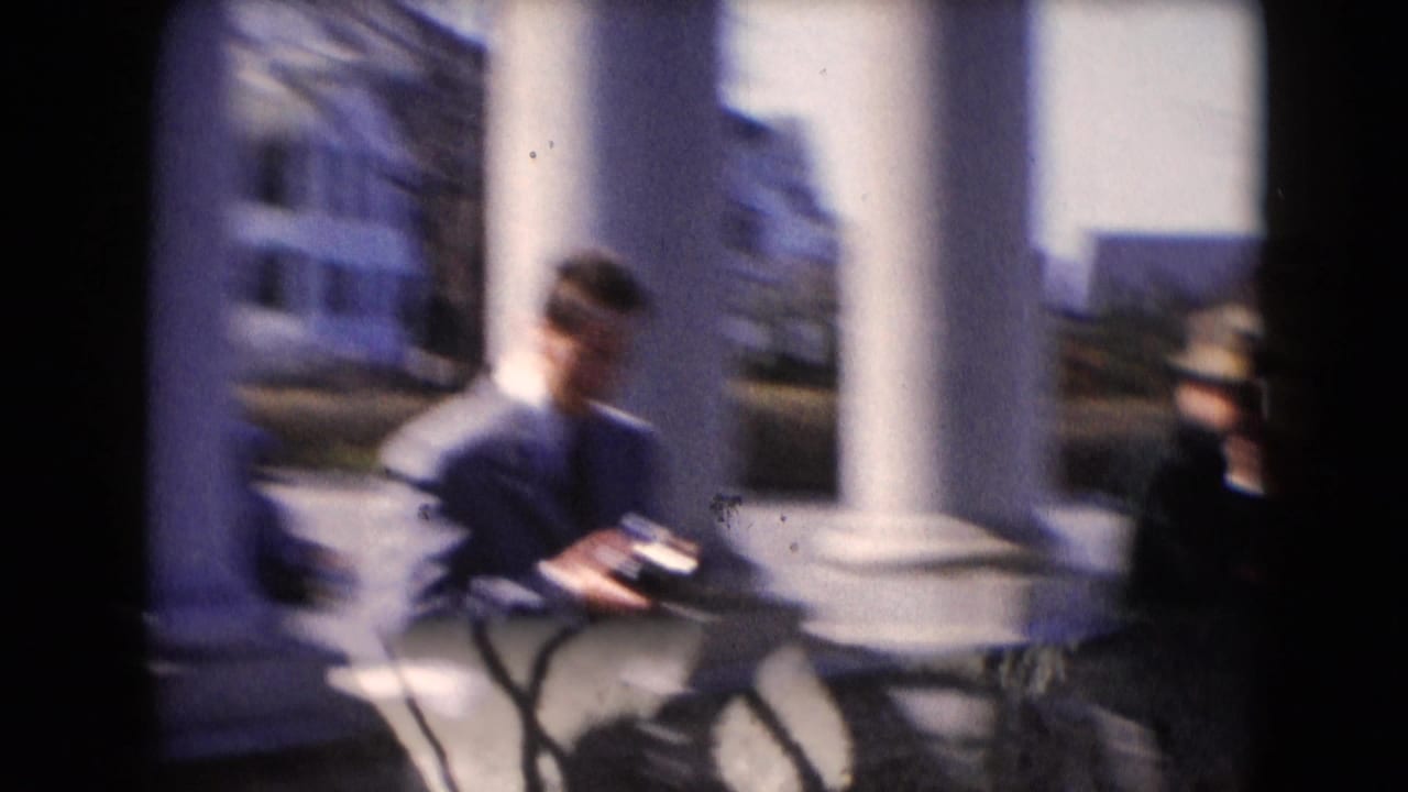 Soft-focused suited man standing near white columns while holding a camera.