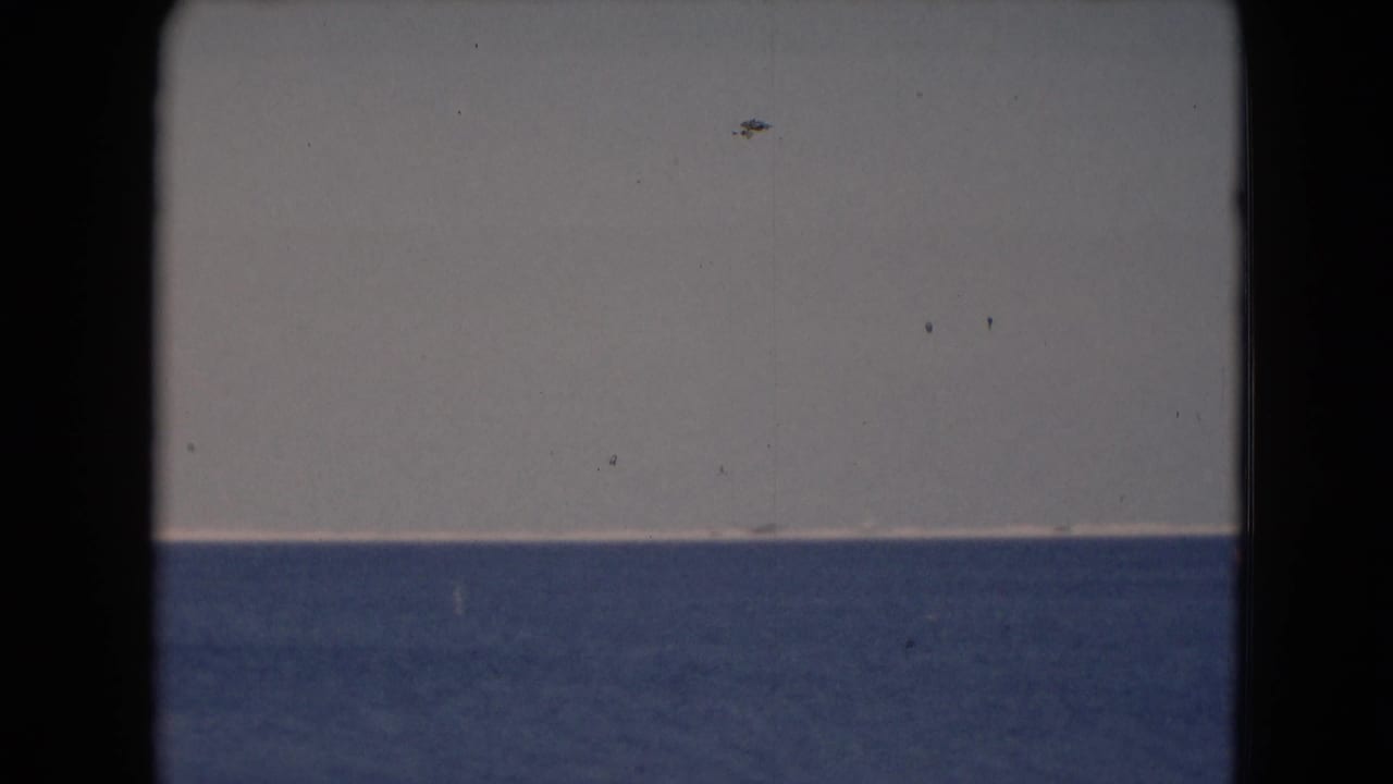 1940s 8mm home-movie frame showing a simple sea horizon, pale sky, and faint distant marks on or above the water.