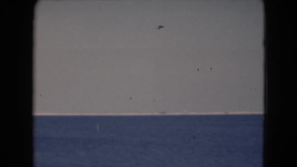 1940s 8mm home-movie frame showing a simple sea horizon, pale sky, and faint distant marks on or above the water.