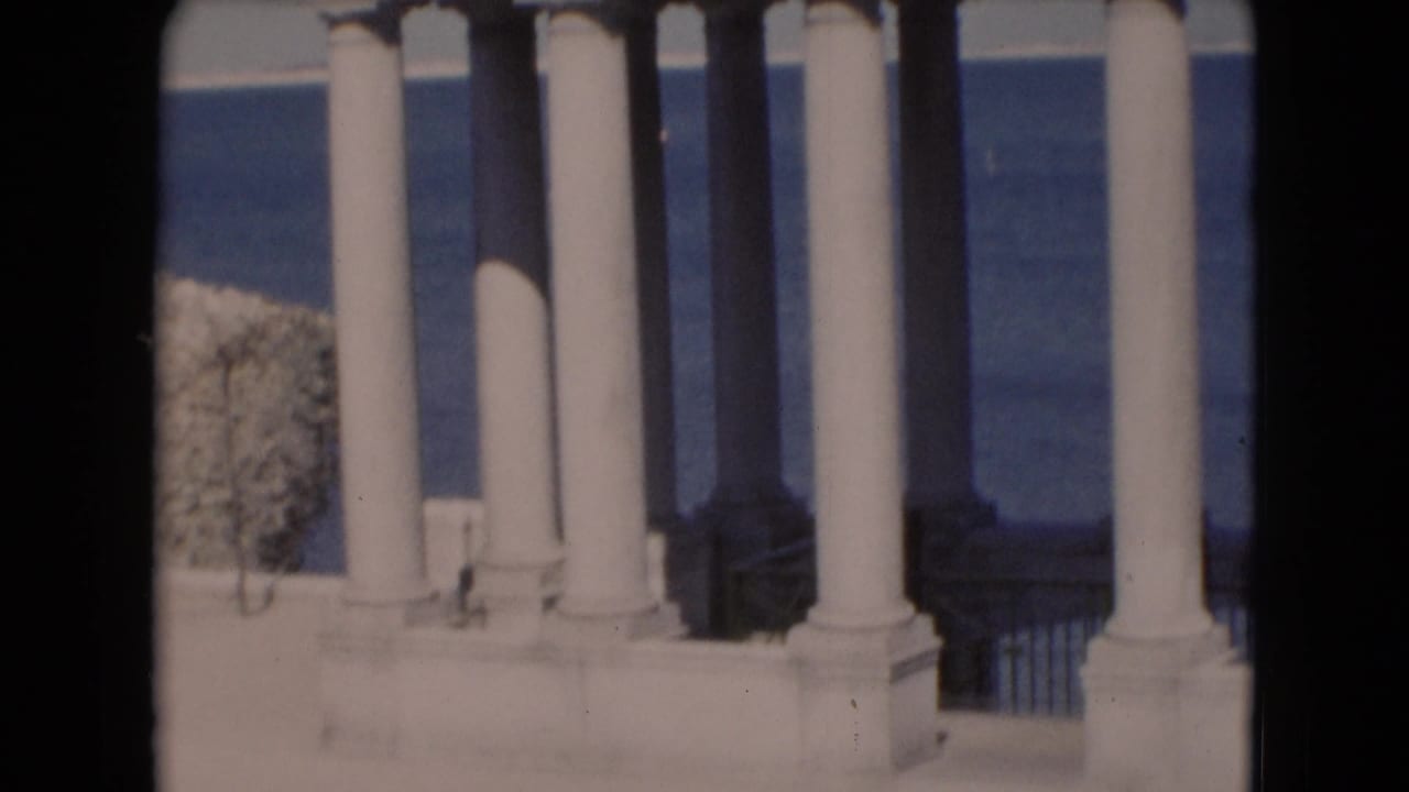 A closer 1940s home-movie frame shows tall white columns, a railing, and water beyond the structure.