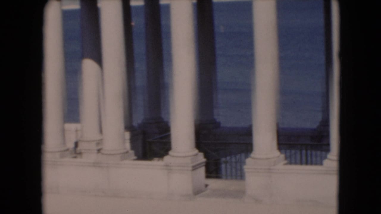 White classical columns and railing with blue water beyond, framed by dark film edges.