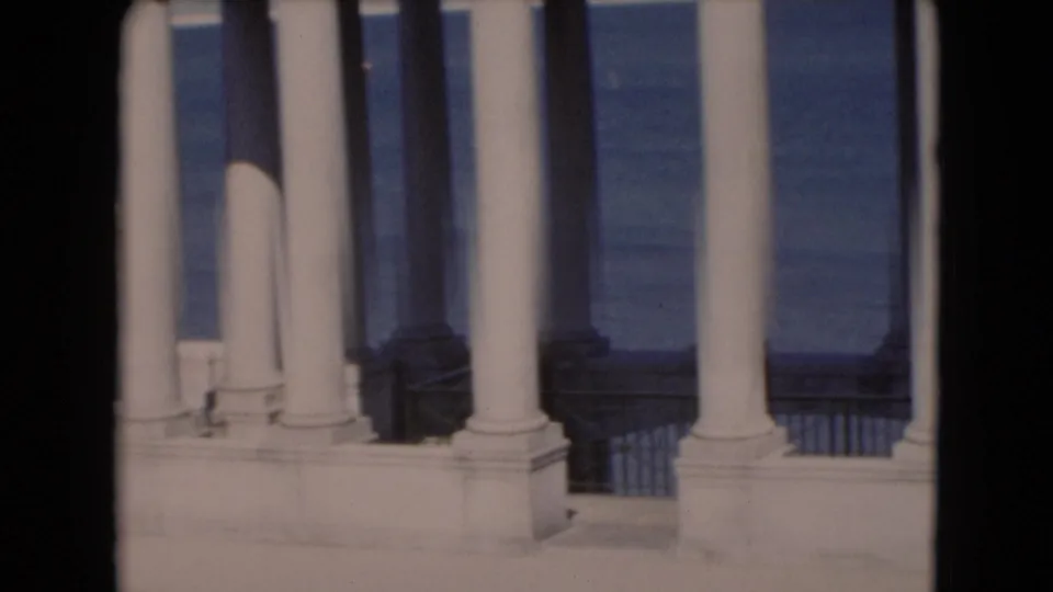 1940s 8mm frame showing white columns and a pale waterfront railing with blue water visible through the colonnade.