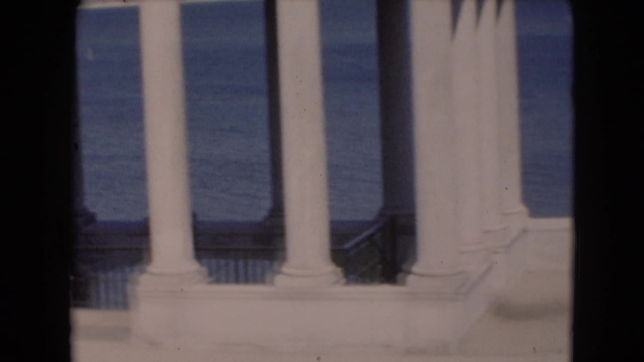 A row of white columns stands before blue water, with a dark vertical film band near center.
