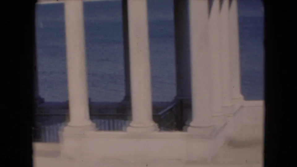 White colonnade seen against blue water in a soft 8mm frame, interrupted by a dark vertical film or gate artifact.