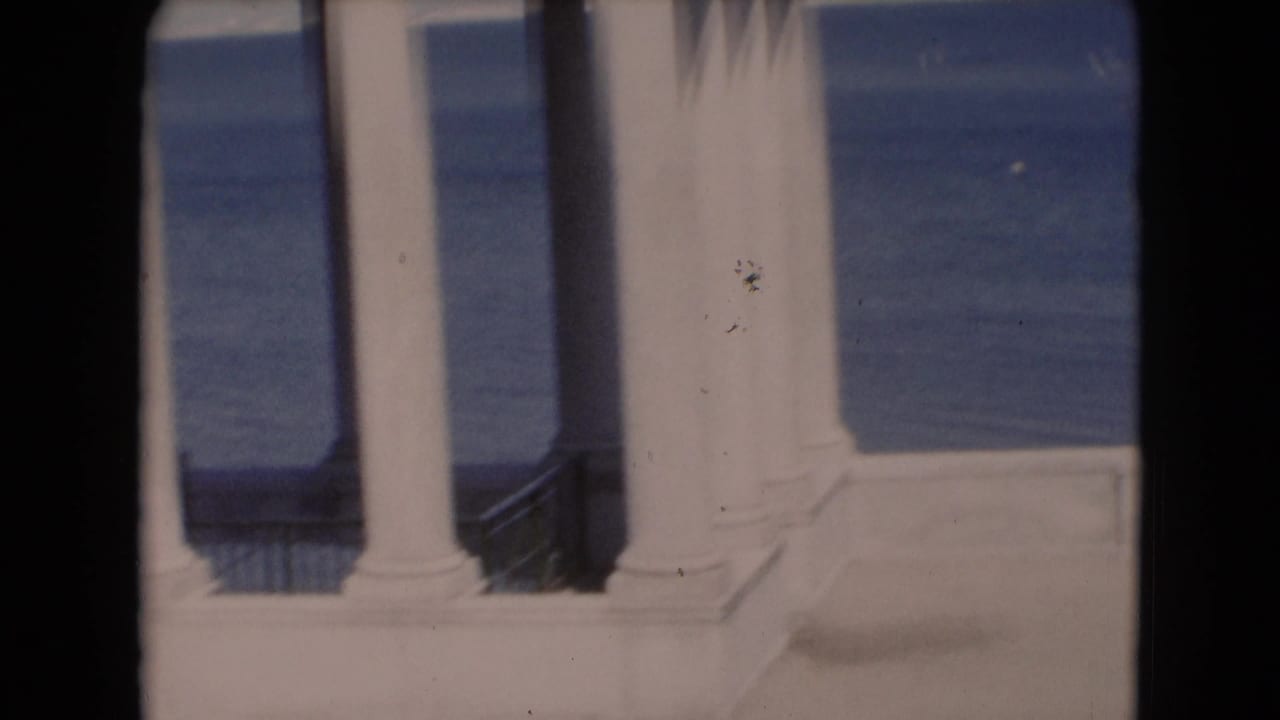 Soft color 8mm view of white waterfront columns and railing, with blue water beyond and visible film-edge shadowing.