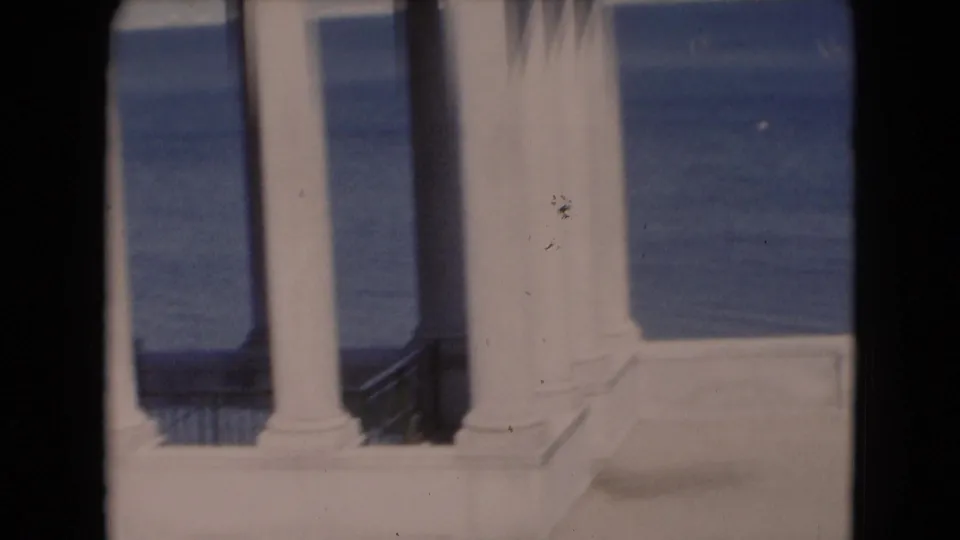 Soft color 8mm view of white waterfront columns and railing, with blue water beyond and visible film-edge shadowing.