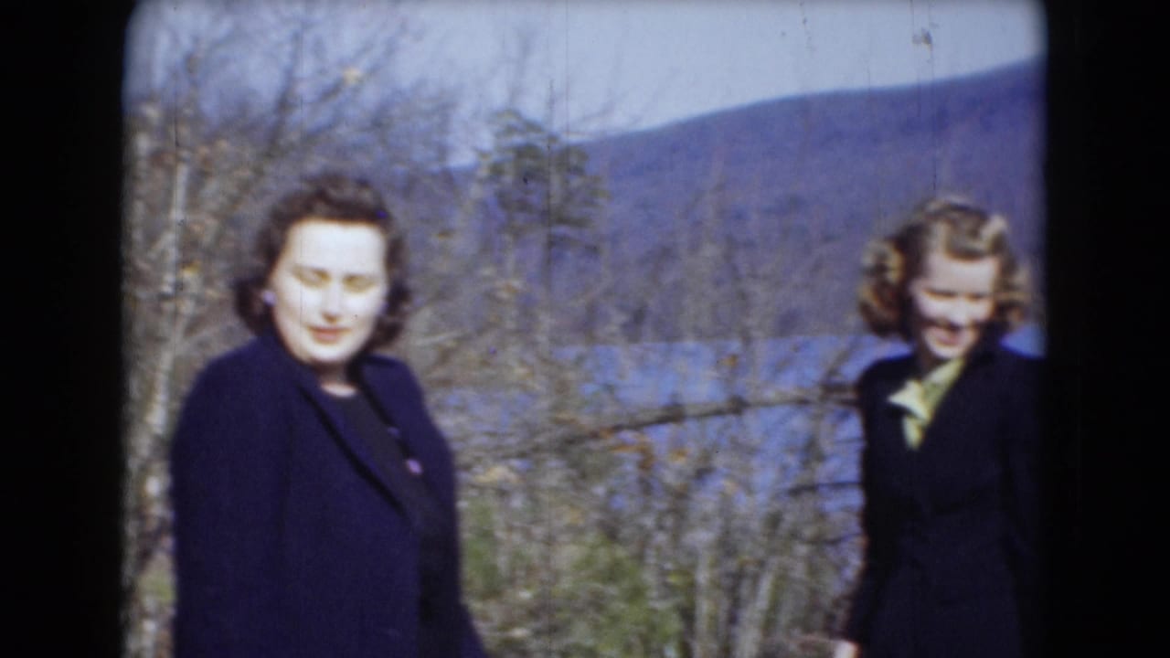 Two women in dark coats pose outdoors near a lake and wooded hills, in archival home-movie footage associated with Middletown, Pennsylvania.