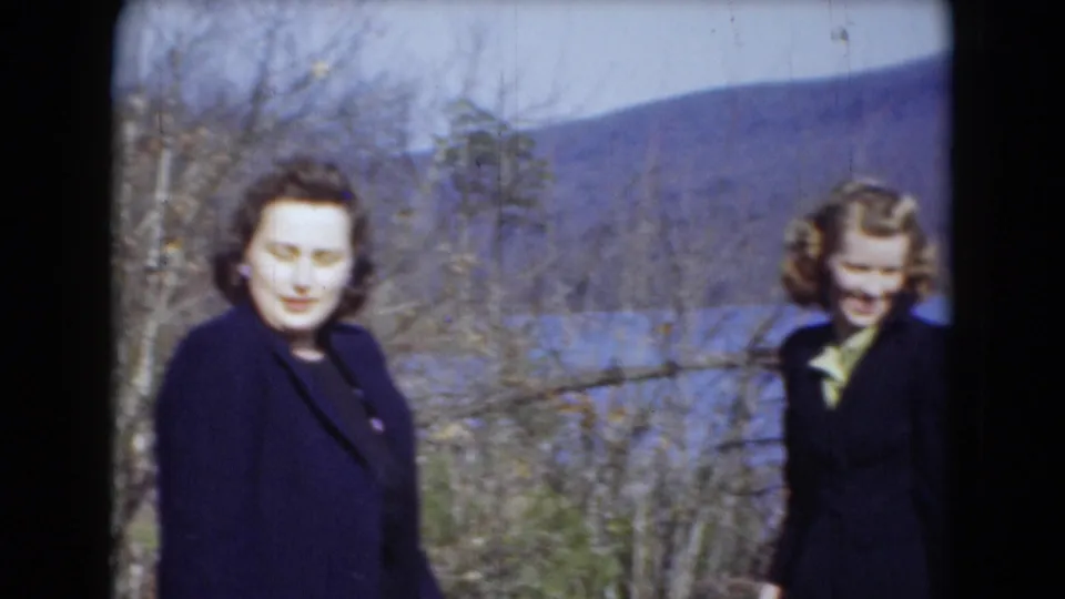 Two women in dark coats pose outdoors near a lake and wooded hills, in archival home-movie footage associated with Middletown, Pennsylvania.