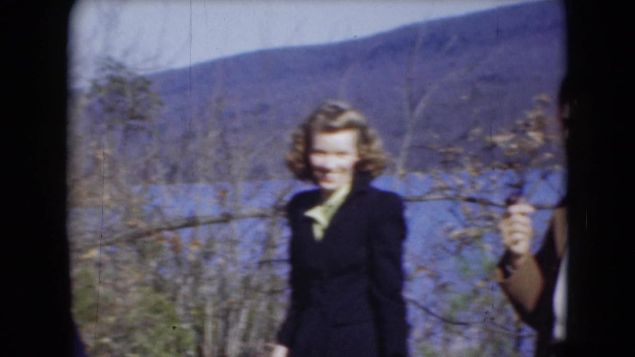 A woman in a dark coat appears near a river in Middletown, Pennsylvania, with bare trees and a wooded hillside behind her.