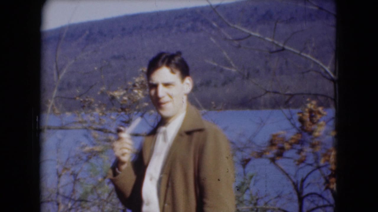 Man in a brown jacket holds a pipe beside a river.