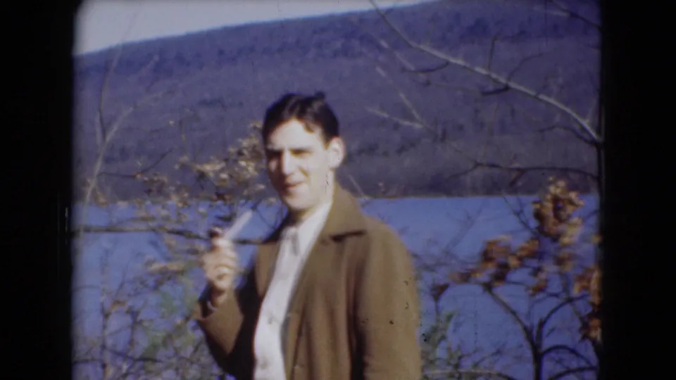 A man in a brown jacket and light shirt holds a pipe outdoors near a river and wooded hillside.