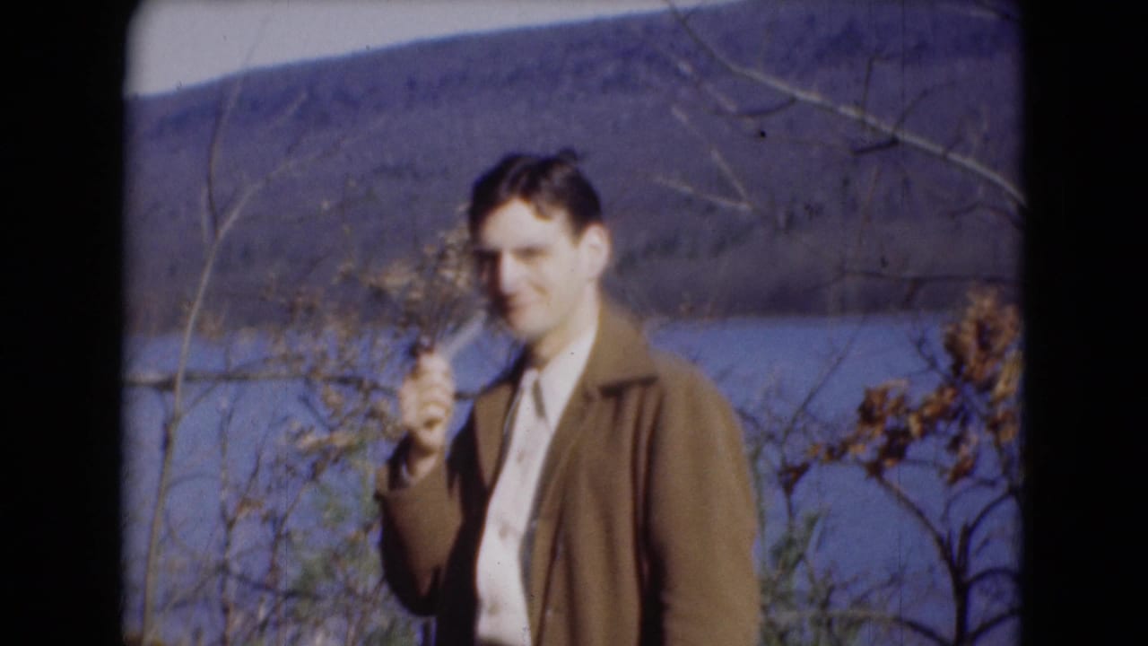 A man with a pipe stands in a brown jacket near the river, framed by bare branches and distant hillside.