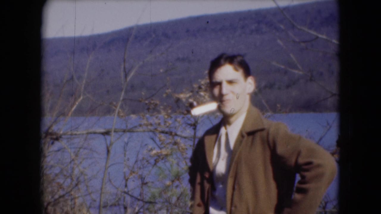 A man in a brown jacket stands by the river with a pipe in his mouth or hand, with bare trees and hillside in the background.