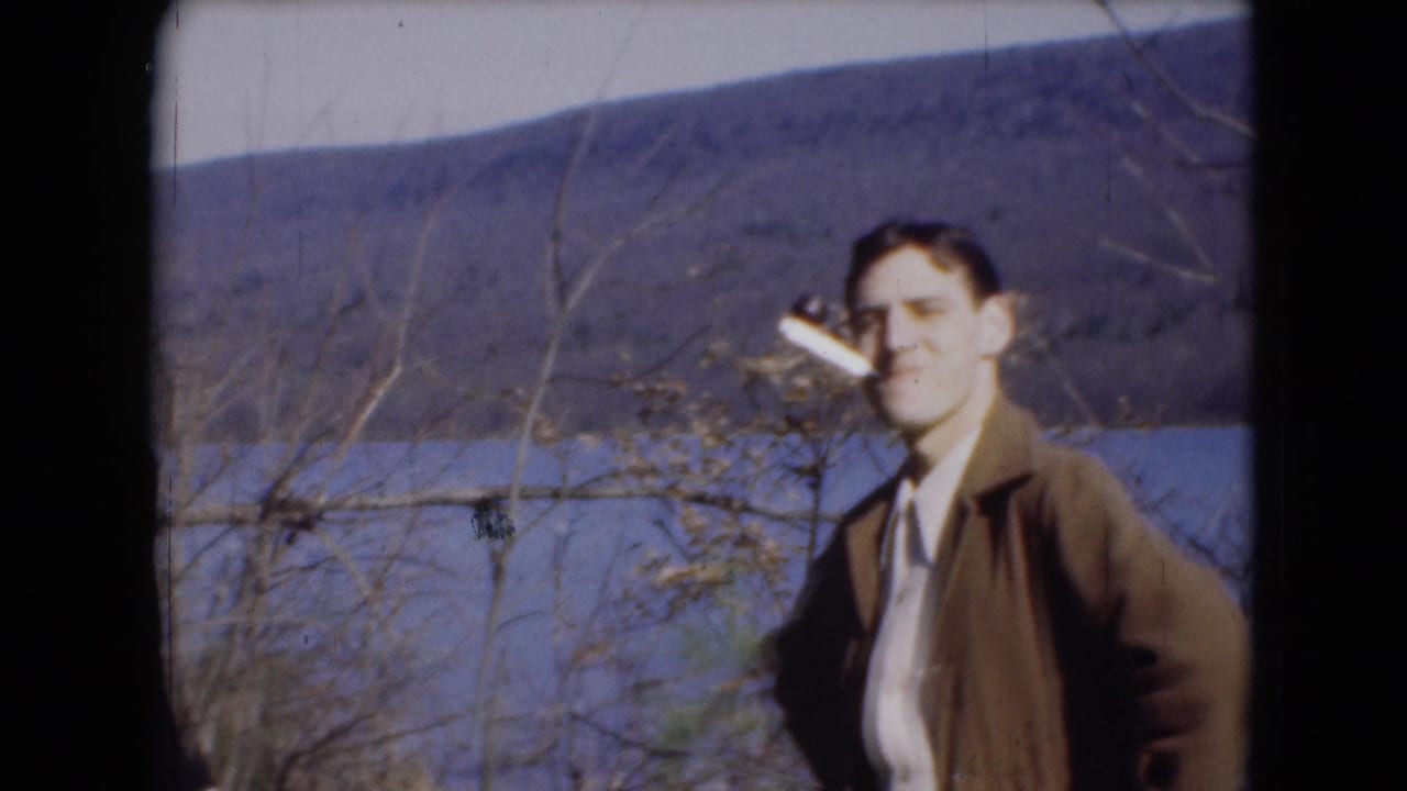 Man in a brown jacket smokes near a lake with hills behind him.