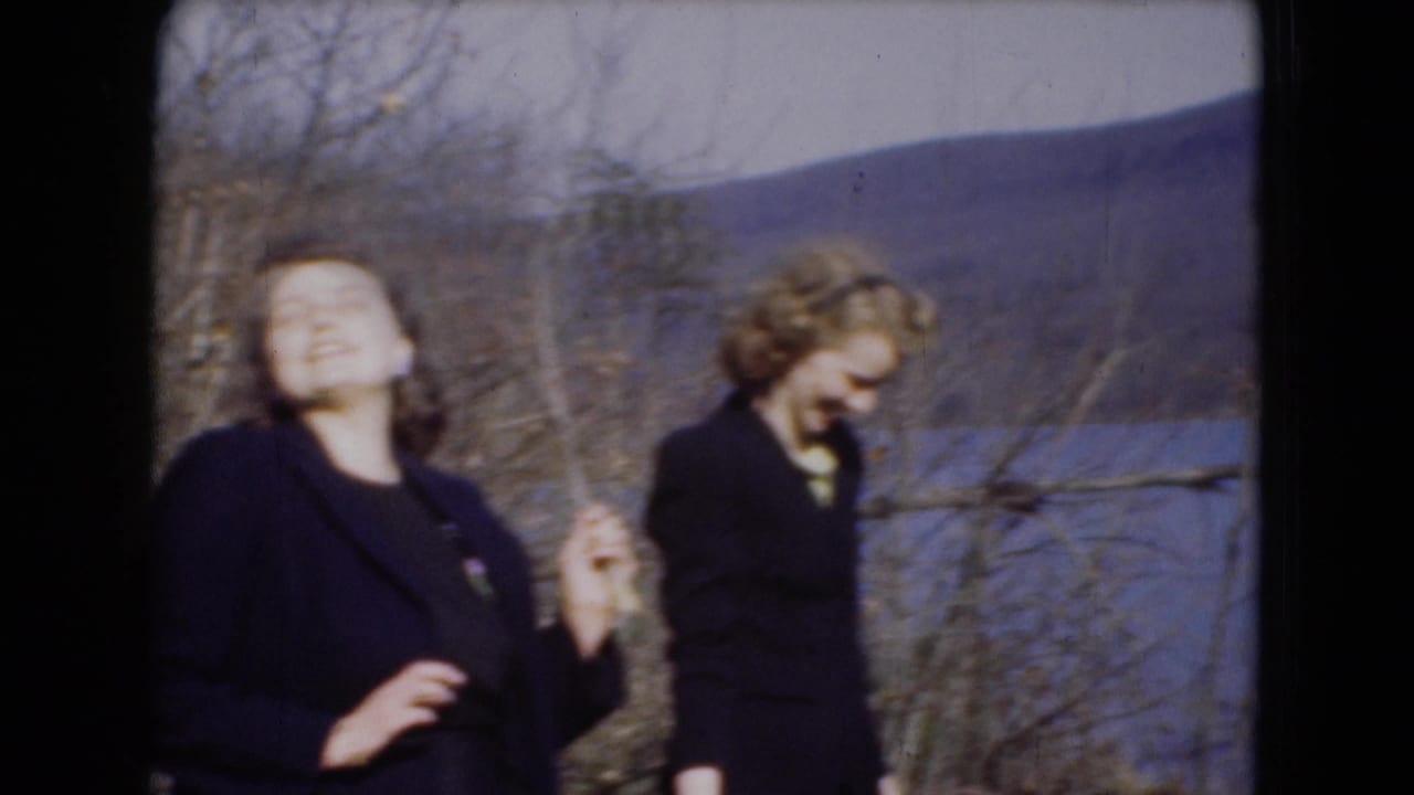 Two women in dark suits stand near a lake, one laughing and one looking down.