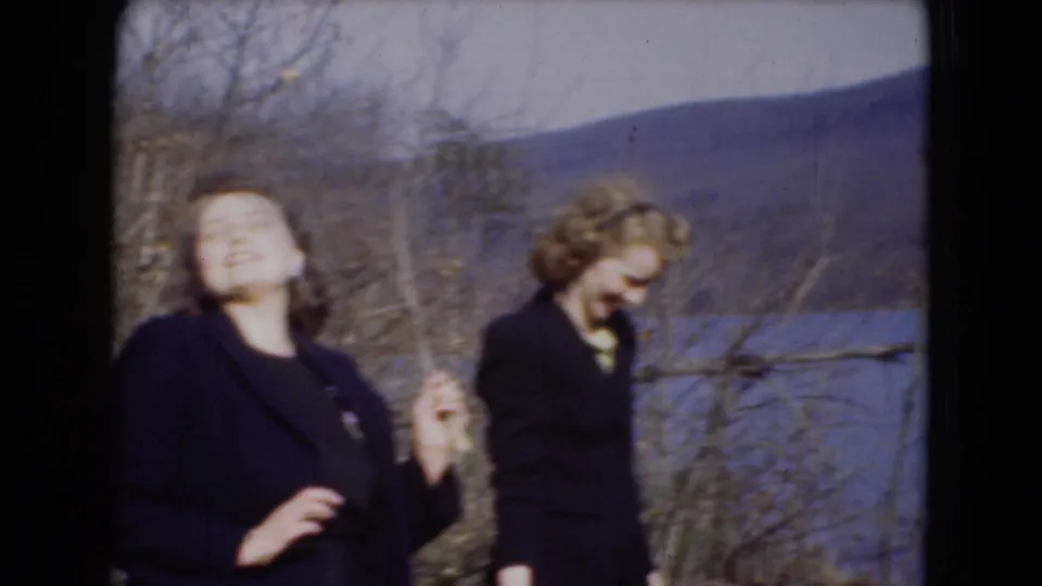 Two women in dark tailored suits appear in 1948 Middletown, Pennsylvania footage near a lake with hills and leafless trees behind them.