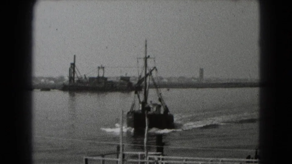 Black-and-white archival frame showing a small working boat or fishing boat passing dock structures with a distant shoreline behind it.