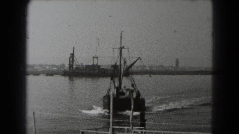 Archival black-and-white frame of a boat moving away near dock posts, with a bird crossing over the water and distant structures behind.