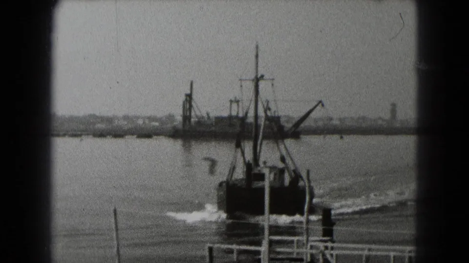Black-and-white 8mm frame of a boat near dock posts, leaving a visible wake across calm waterfront water.