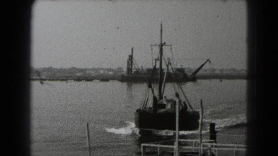 Black-and-white 8mm frame of a small working boat crossing harbor water near Middletown, Pennsylvania, with an industrial vessel beyond it.