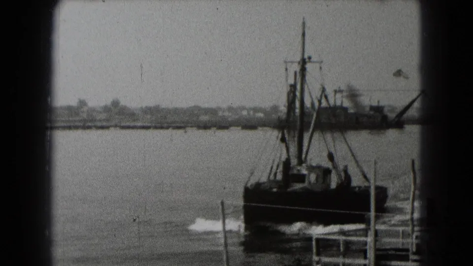 The small harbor boat continues past the camera in black-and-white 8mm footage, with a larger industrial vessel and shoreline behind it.