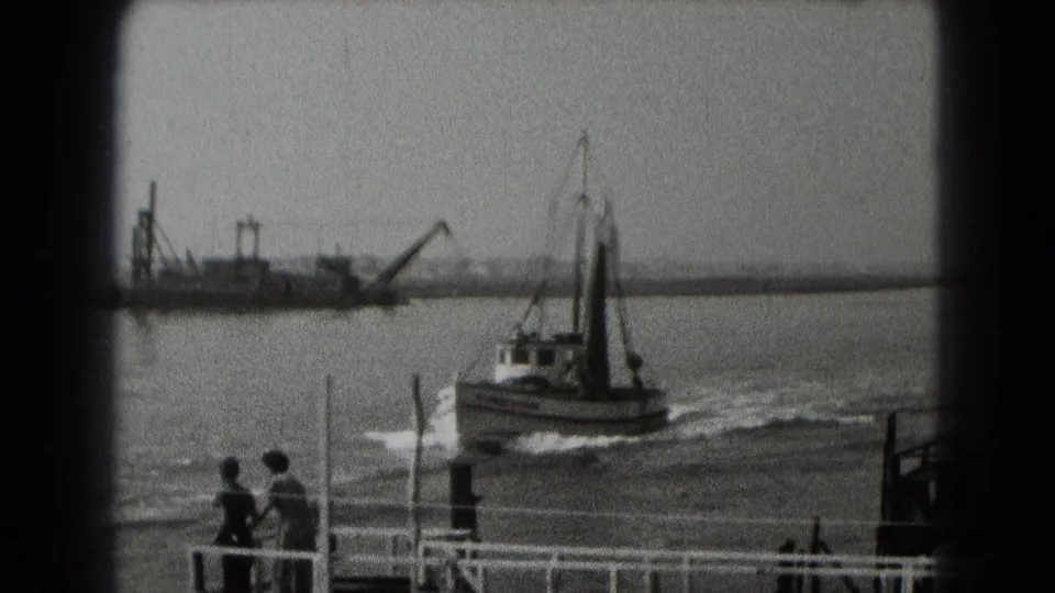 Dockside view of a small boat moving through the harbor, with two distant people watching from the railing in this 1948 home-movie frame.