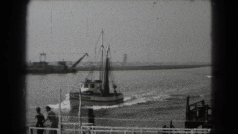 The boat moves farther along the harbor in a soft black-and-white 8mm frame, with two dockside figures visible in the foreground.