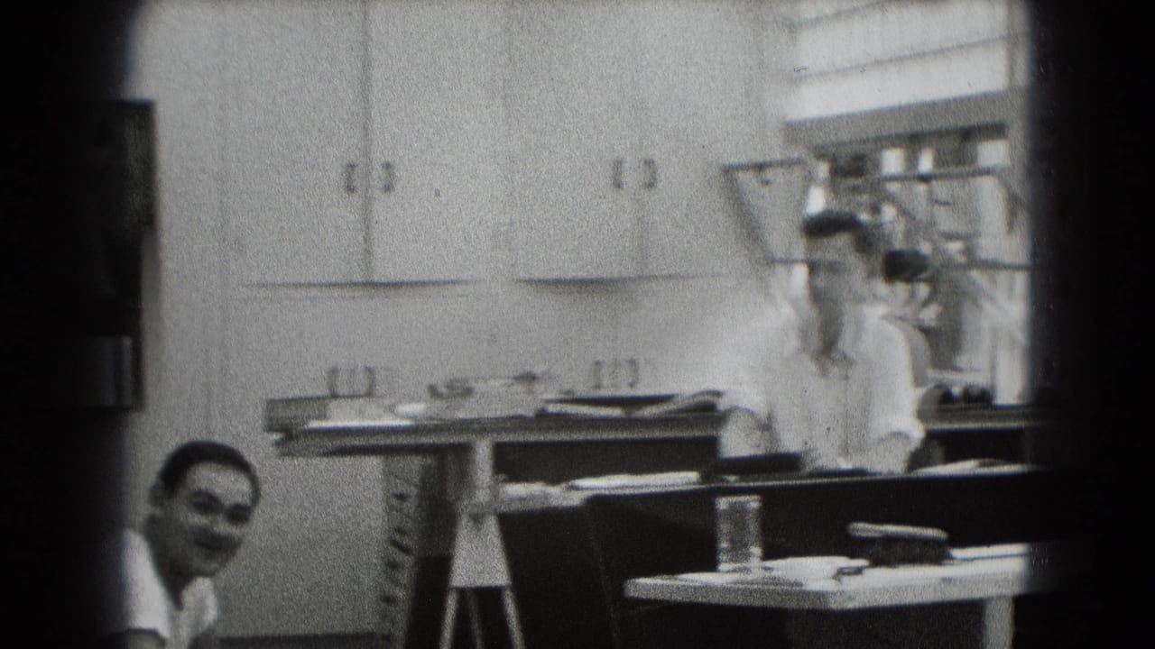 Two adult men in a small room with cabinets, table clutter, and one man smiling near the foreground.