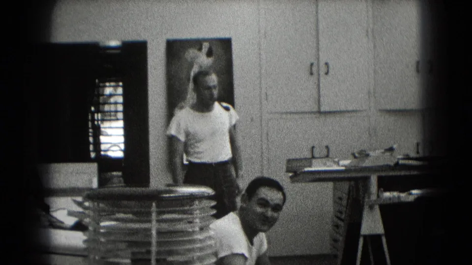 Mid-century 8mm frame from Middletown shows two adult men in a private room with cabinets, a wall picture, and stacked discs or plates.