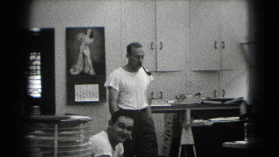 A 1948 Middletown home-movie frame shows an adult man smoking a pipe beside a table, with a second man smiling toward camera.