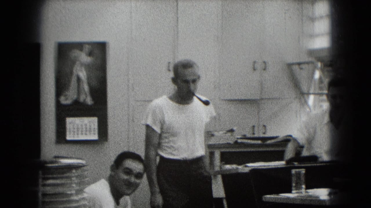 Man with a pipe stands in an office while another man sits nearby.