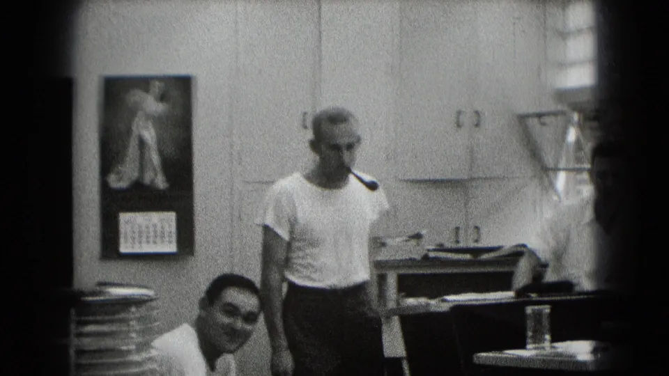 In a Middletown, Pennsylvania home-movie frame, a man with a pipe stands near desks, cabinets, a calendar, and another seated man.