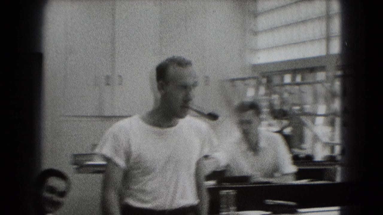 A man with a pipe crosses an office-like interior with wall cabinets, blinds, and another adult man partly visible beside him.