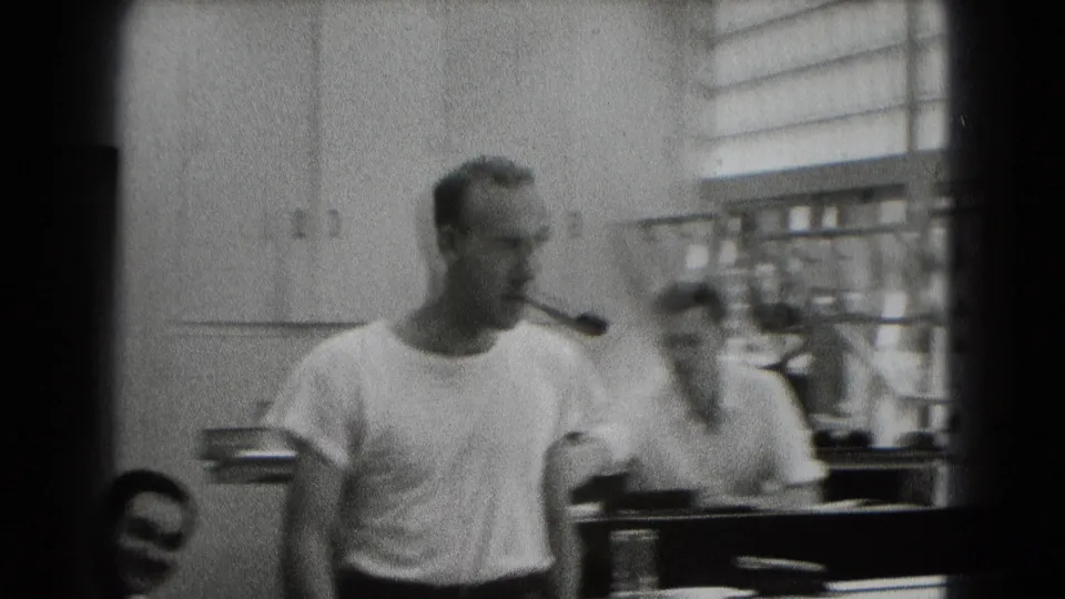 A man with a pipe crosses an office-like interior with wall cabinets, blinds, and another adult man partly visible beside him.