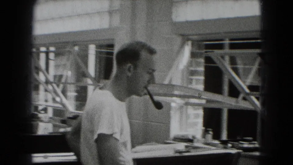 The man with a pipe is seen in profile near a work surface, with blinds, railings or racks, and office items behind him.