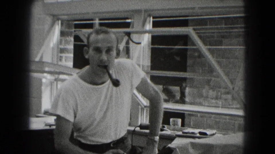 A man with a pipe faces toward the camera in the Middletown interior, with desks, shelves, blinds, and small office objects visible around him.