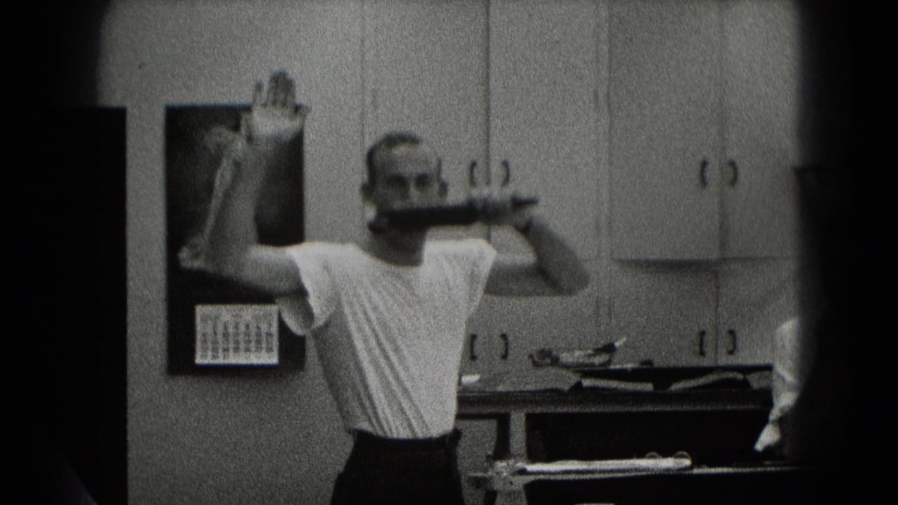 Man in a white T-shirt holds a pipe near wall cabinets and a calendar.