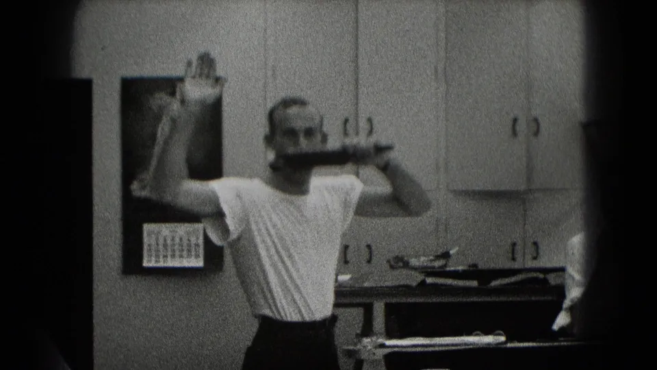 In Middletown, Pennsylvania home-movie footage, a man in a white T-shirt holds a pipe and gestures near cabinets and a wall calendar.
