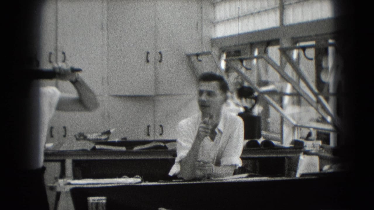 Two adult men are visible in a cabinet-lined Middletown interior, with one seated at a counter and another partly cropped while holding a pipe.