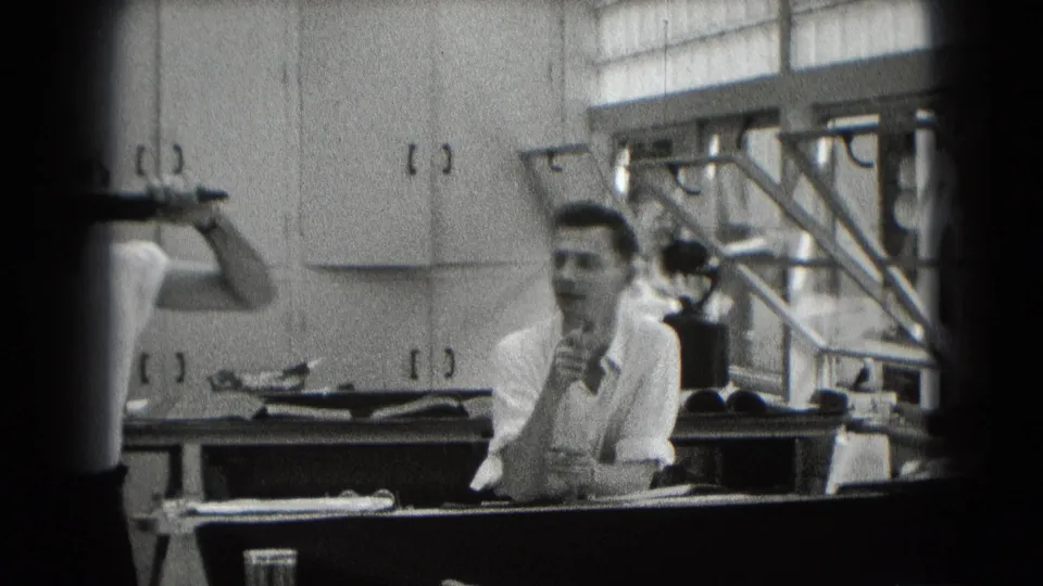 Two adult men are visible in a cabinet-lined Middletown interior, with one seated at a counter and another partly cropped while holding a pipe.