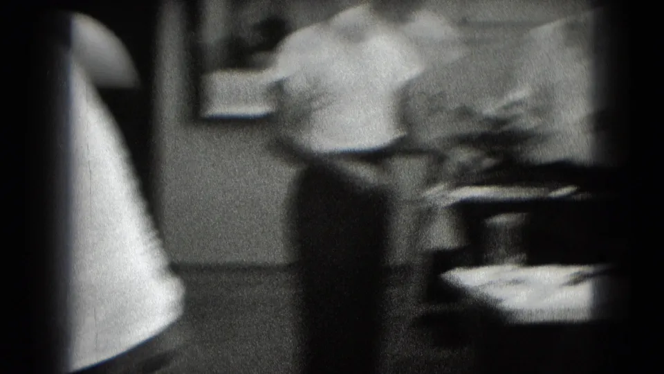 Grainy 8mm frame from a private interior, likely in Middletown, Pennsylvania, with a blurred man near a worktable.