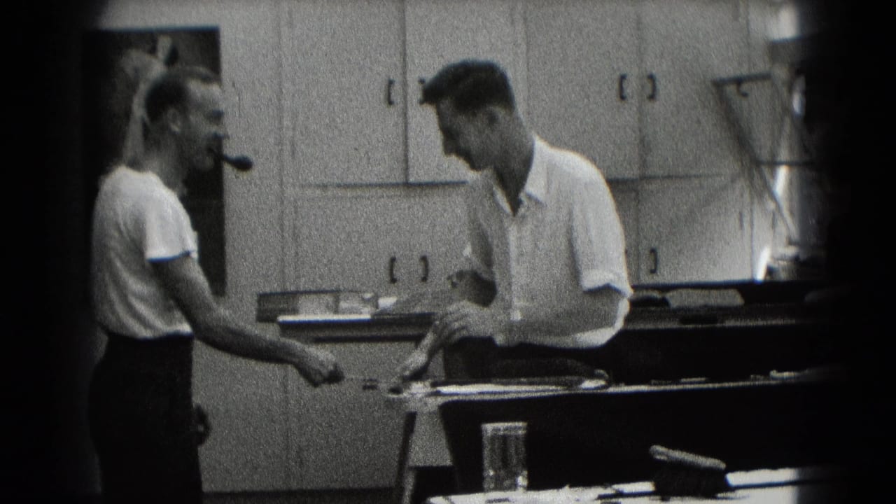 Two adult men in a Middletown workplace interior, one with a pipe, converse beside a workbench and wall cabinets.