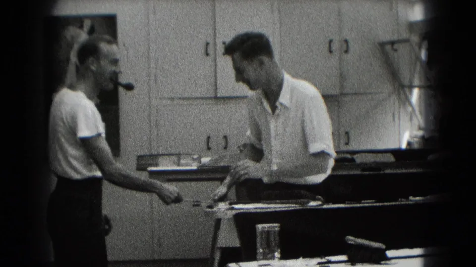 Two adult men in a Middletown workplace interior, one with a pipe, converse beside a workbench and wall cabinets.