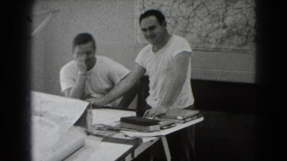 Two adult men in white shirts sit or lean beside a table with rolled plans and books in a private workshop or office.