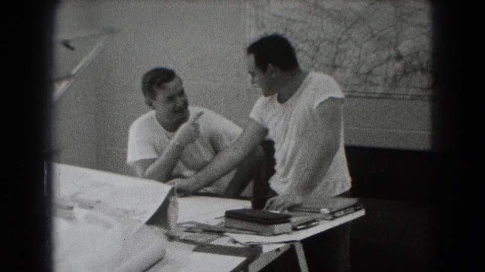 Two men converse closely at a worktable in a grainy 8mm Middletown interior, with papers and a wall map visible.