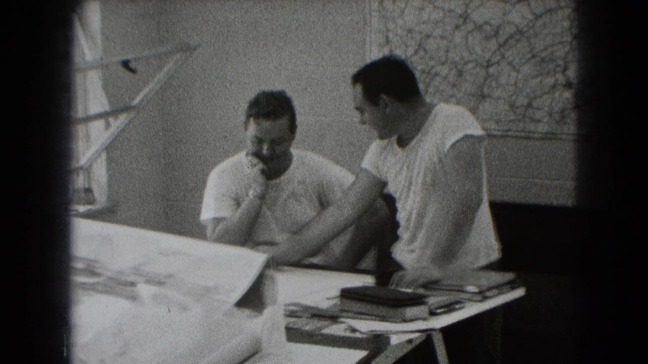 Two men sit at a table reviewing large plans near a window.