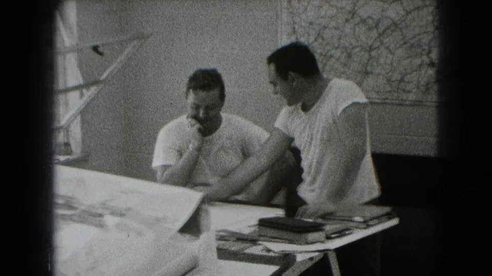 Black-and-white 8mm frame of two men in a Middletown, Pennsylvania interior reviewing large plans on a table.
